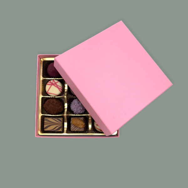 Rosa Pralinenbox quadratisch aus festem Karton. Deckel leicht geöffnet und schräg darüber gelegt, dass man einen Teil der Pralinen sieht.