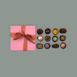 Rosa Pralinenbox für 12 Pralinen in quadratischer Form. Mit rosa Schleife und kleiner braunen Schleife. Daneben aufgereiht zwölf verschiedene Pralinen