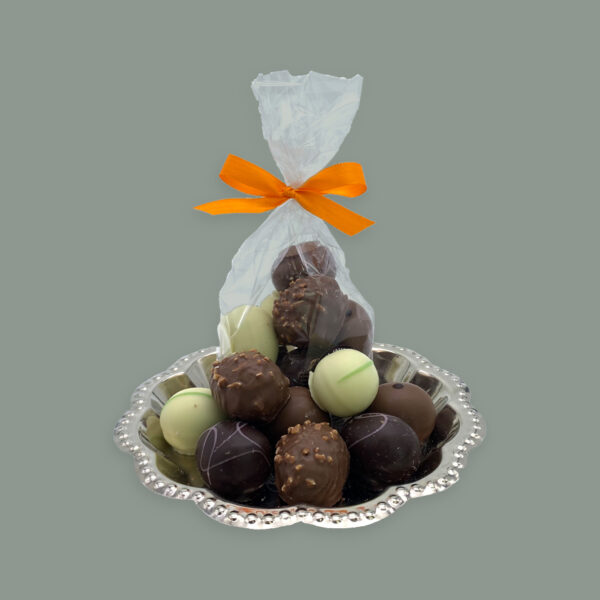 Coppeneur Pralinen in Ostereiform. In diversen Geschmacksrichtungen. Umhüllt von weißer Schokolade, Zartbitterschokolade und Vollmilchschokolade. Hier dargestellt auf kleinem runden silbernem Tablett. Im Vordergrund unverpackt dahinter in einer durchsichtigen Tüte mit orangener Schleife.