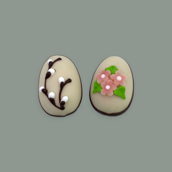 Zwei Marzipan Ostereier halb getaucht in Zartbitterschokolade. links verziert mit Zartbitterschokolade und weißer Schokolade, als Weidenkätzchenstrauch. Recht verziert mit Blumen aus Zuckerguss.