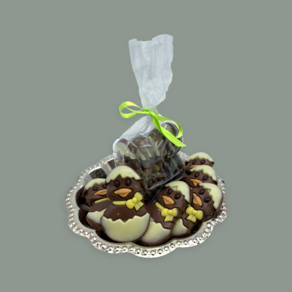 Schokoladen Flachfigur: Kleine Küken in Eierschale. Vollmilchschokolade verziert mit Weißer Schokolade. Hier dargestellt auf kleinem runden silbernem Tablett. Im Vordergrund unverpackt dahinter in einer durchsichtigen Tüte mit grüner Schleife.