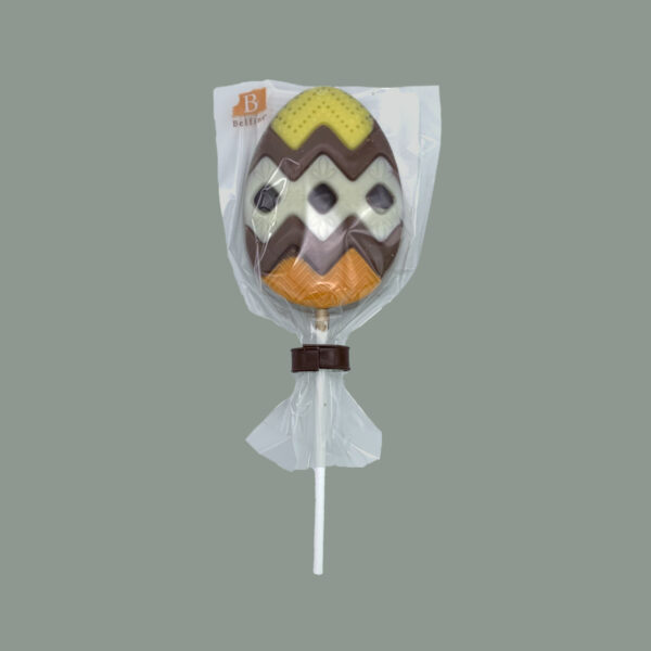 Lolly in Osterei Form aus Vollmilchschokolade grafisch verziert mit gefärbter weißer Schokolade. Verpackt in durchsichtigem Tütchen.