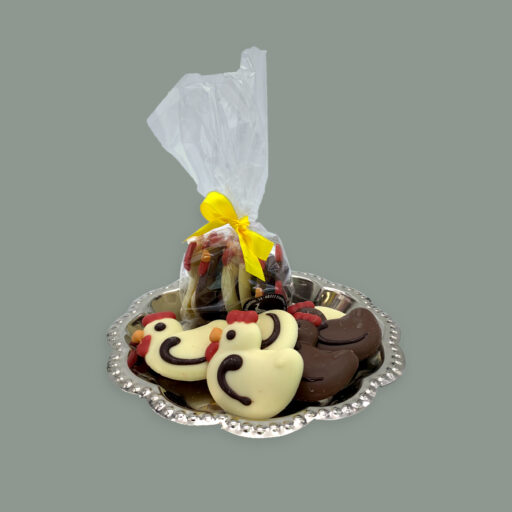 Schokoladen Flachfiguren aus weißer Schokolade und Vollmilchschokolade, dekoriert als kleine Hennen. Hier dargestellt auf kleinem runden silbernem Tablett. Im Vordergrund unverpackt dahinter in einer durchsichtigen Tüte mit gelber Schleife.