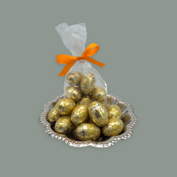Ostereier Von Venchi in goldenem Stanniol aus Nougat. Hier dargestellt auf kleinem runden silbernem Tablett. Im Vordergrund unverpackt dahinter in einer durchsichtigen Tüte mit orangener Schleife.