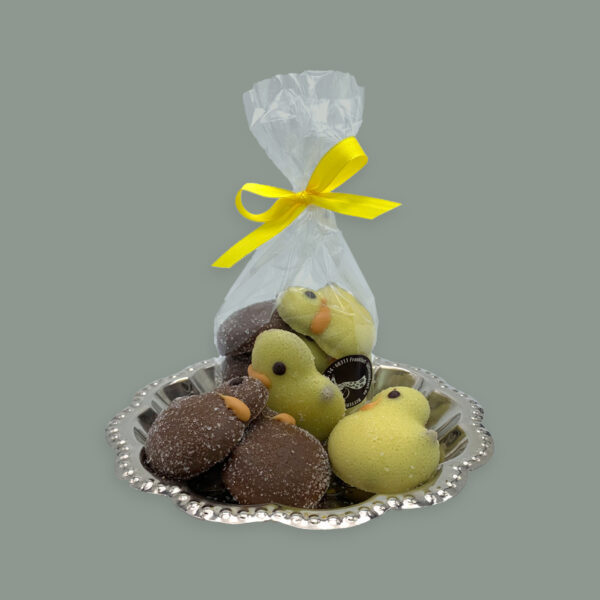 Küken aus weißer Schokolade und Vollmilchschokolade gewälzt in Zucker. Gefüllt mit Nougat. Hier dargestellt auf kleinem runden silbernem Tablett. Im Vordergrund unverpackt dahinter in einer durchsichtigen Tüte mit gelber Schleife.
