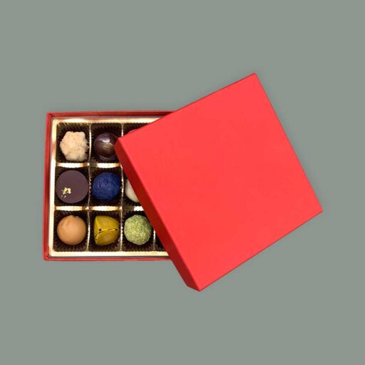 Pralinenschachtel quadratisch in Rot. Der Deckel liegt nur halb auf der Schachtel, sodass man ein Blick im die Schachten gefüllt mit verschiedenen Pralinen sieht.